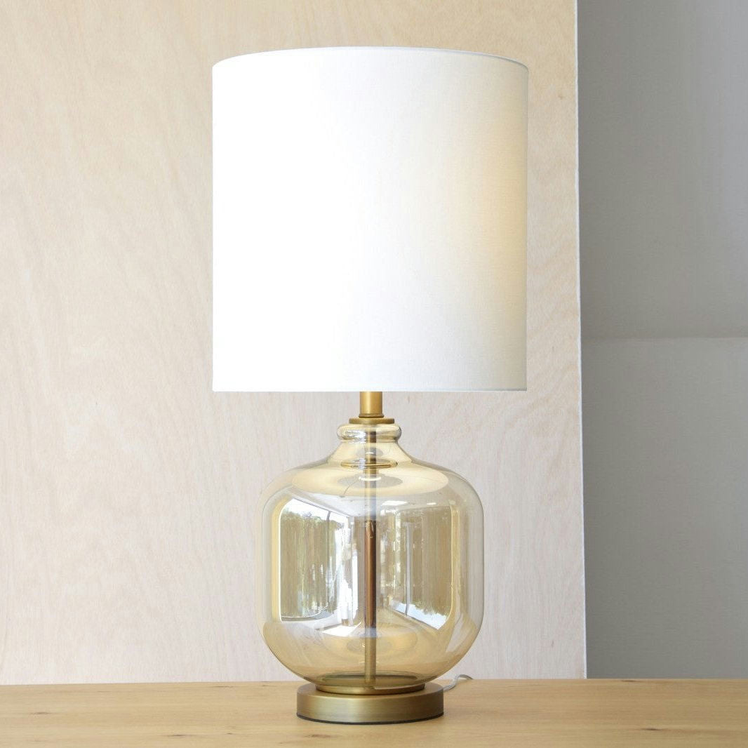 La Lámpara de mesa Bela en Vidrio Ámbar con Pantalla se asienta elegantemente sobre una superficie de madera, incorporando un diseño exclusivo frente a la pared luminosa.