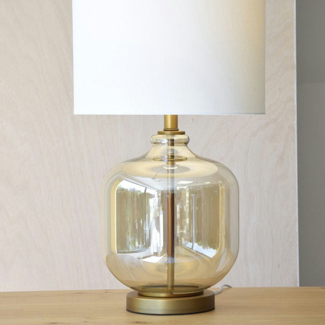 La lámpara de mesa Bela en vidrio ámbar presenta una base redonda transparente y acabado dorado. Su pantalla beige se eleva con elegancia, ideal para interiores atractivos.