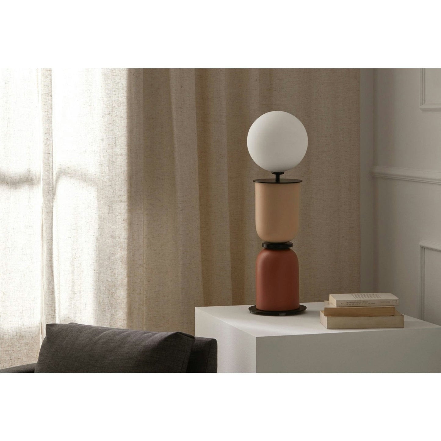 La lámpara de mesa Copo con bombilla esférica reposa sobre un pedestal geométrico junto a unos libros apilados sobre una mesa blanca. Esta escena de diseño minimalista se completa con un sillón gris y unas cortinas beige de fondo.