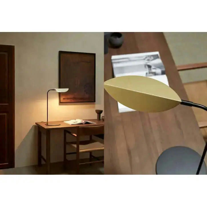 La elegante lámpara de mesa Ficus ilumina una mesa de madera con su diseño moderno. Un primer plano a la derecha muestra su hermosa forma de hoja, mientras que una pintura encantadora agrega encanto a esta elegante escena interior.