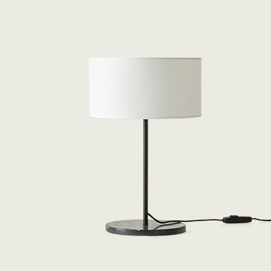 La Lámpara de mesa Hedra, con su pantalla cilíndrica blanca sobre un delgado tallo negro y base circular, cuenta con un cable y un interruptor negros. Rezuma diseño contemporáneo a la vez que proporciona una iluminación acogedora.