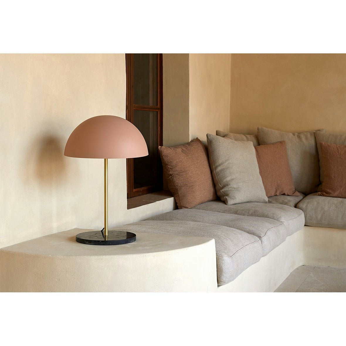 Un acogedor rincón de la sala de estar cuenta con un sofá modular beige con cojines neutros. La lámpara de mesa Hedra marrón con forma de cúpula y soporte dorado sobre una mesa auxiliar semicircular agrega un elemento de iluminación acogedora que combina perfectamente con el diseño contemporáneo.