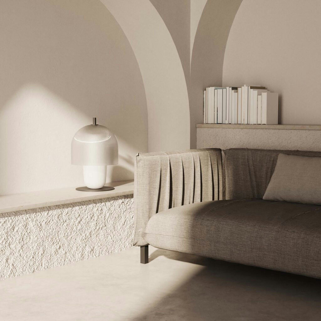 Una sala de estar minimalista presenta la simplicidad escandinava con un sofá gris plisado, libros apilados y la lámpara de mesa Holm que proporciona una iluminación ambiental cálida desde una repisa texturizada contra una pared arqueada.