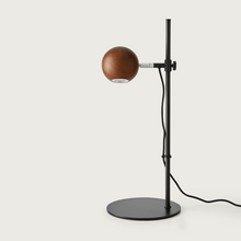  Lámpara de mesa Lita con pantalla esférica de madera, esbelto soporte de metal negro y base circular sobre fondo blanco, de diseño minimalista.