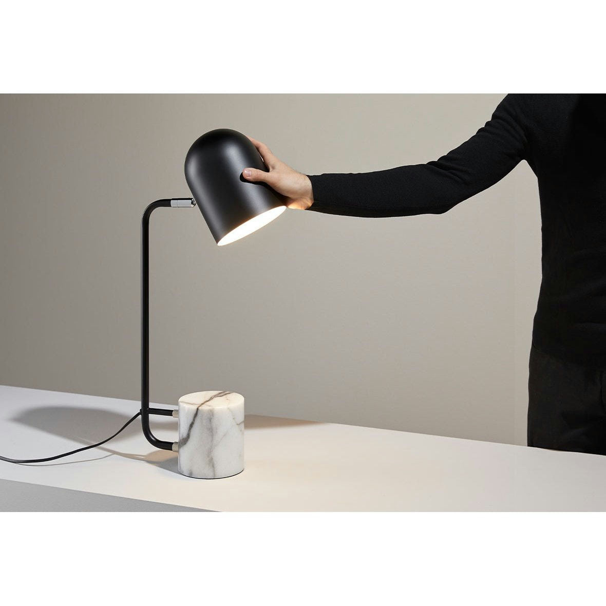 Alguien ajusta cuidadosamente la Lámpara de mesa Luca, mostrando un diseño contemporáneo con un elegante acabado negro y una elegante base de mármol blanco, que complementa perfectamente la limpia superficie blanca.