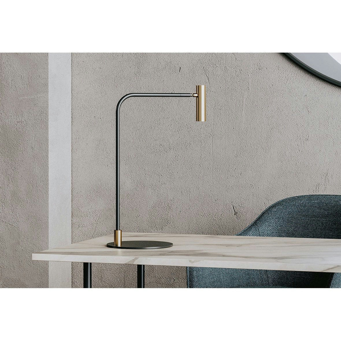 La lámpara de mesa Maho cuenta con un elegante brazo negro y detalles dorados con iluminación LED, que reposa sobre una mesa clara. Complementa elegantemente la pared gris texturizada y se ubica junto a una silla azul parcialmente visible.