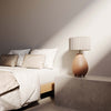 Un dormitorio minimalista con una cama blanca, almohadas beige y una exclusiva Lámpara de mesa Obrie sobre una mesita de noche gris, bañada por la luz natural del sol.