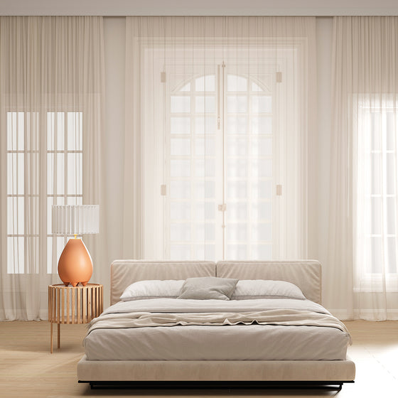 Un dormitorio minimalista con una gran cama centrada frente a una ventana luminosa enmarcada por cortinas transparentes, en tonos neutros y una mesa auxiliar con una Lámpara de mesa Obrie (Cerámica o Metal).