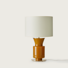  Una moderna lámpara de mesa Ponn con pantalla cilíndrica blanca y base brillante de vidrio ámbar, aislada sobre un fondo blanco.