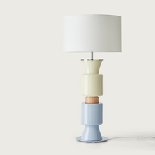  Lámpara de mesa Ponn Ponn con pantalla blanca sobre una base cerámica apilada multicolor en tonos crema, marrón y azul, contra un fondo blanco.