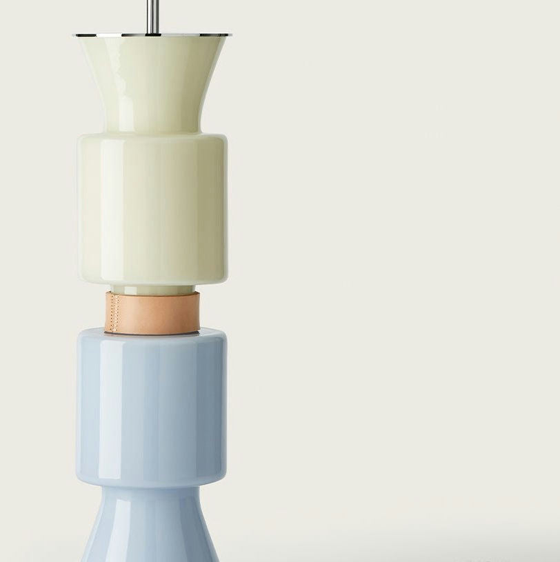 Una pila de tres jarrones de cerámica que muestra un diseño innovador con un amarillo pálido en la parte superior, una banda de cuero en el medio y azul claro en la parte inferior, sobre un fondo gris claro. Producto: Lámpara de mesa Ponn Ponn.