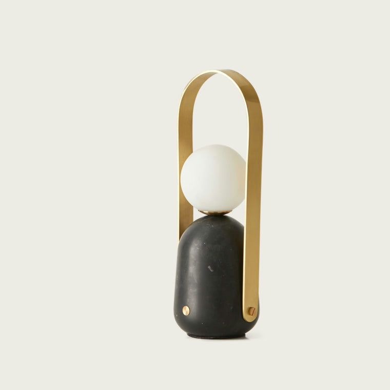 La lámpara de mesa Portátil Nano presenta un diseño moderno con una bombilla esférica blanca sobre una base negra y un mango dorado en forma de arco, realzando a la perfección cualquier espacio con su estilo elegante y encarnando el diseño moderno.