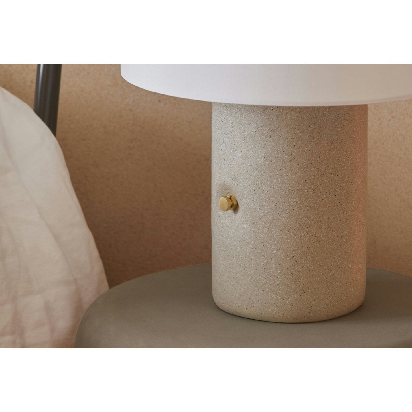Un primer plano de la Lámpara de mesa Sand revela su base cilíndrica con textura de piedra Terrazzo Gris y su pequeño pomo de latón. Con un sistema regulable para una iluminación perfecta, el fondo incluye una almohada y una pared de color neutro.