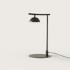 Lámpara de escritorio minimalista de color negro, con cuello curvo y pantalla en forma de cúpula, colocada sobre una superficie blanca con un cordón visible. Esta Lámpara de mesa Tana presenta elementos de diseño moderno para una apariencia atractiva.