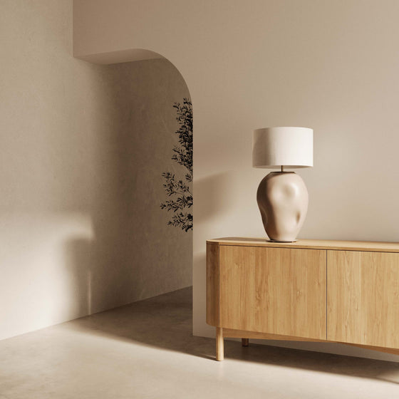 Un rincón de habitación minimalista con un aparador de madera, sobre el que descansa una Lámpara de mesa Ural que asemeja una cabeza humana, junto a una pared curva con un sutil diseño floral.