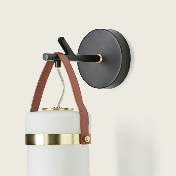 La lámpara de pared Almon presenta correas de cuero que aseguran un diseño cilíndrico blanco y dorado, elaborado con materiales de alta calidad.