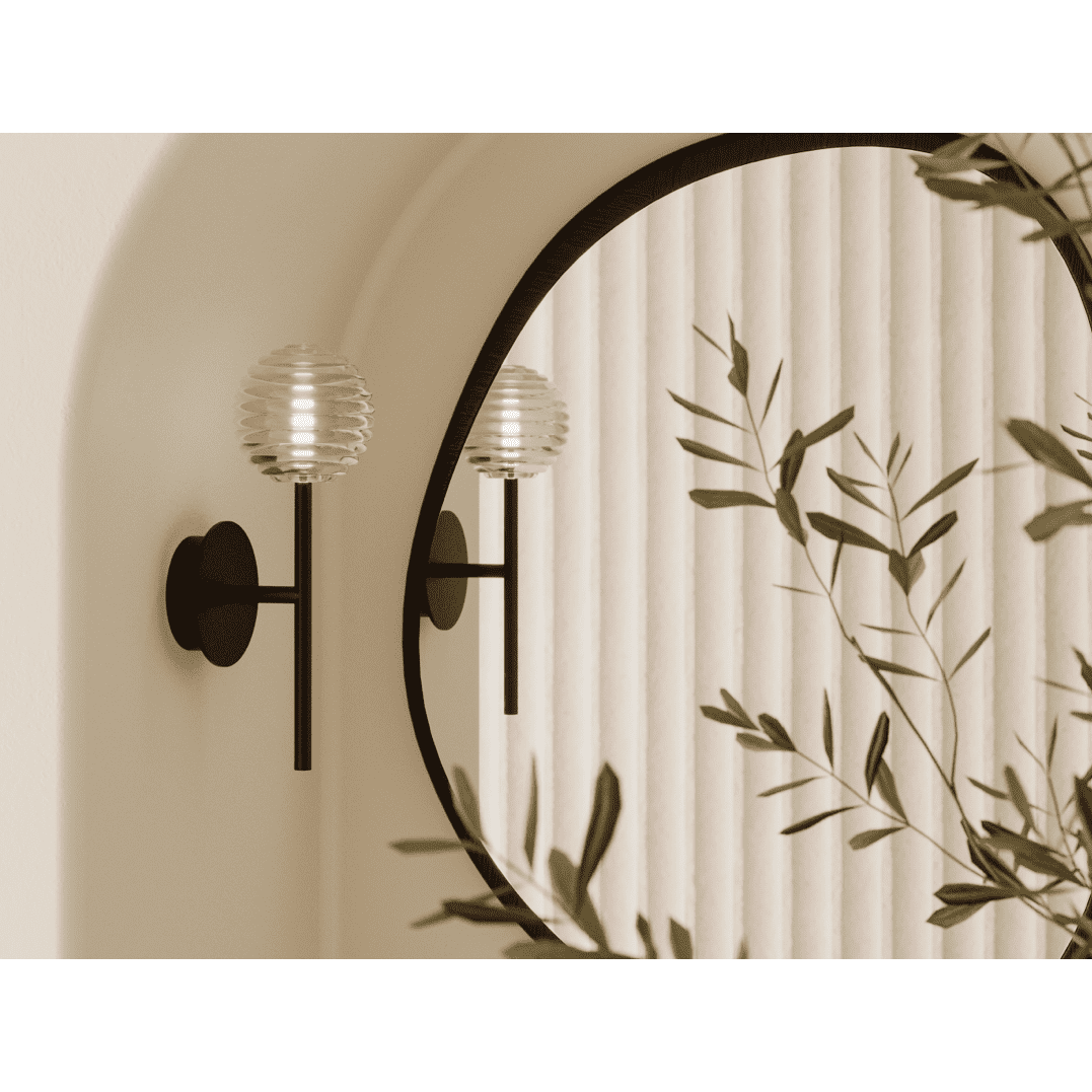 Un espejo redondo refleja un aplique negro con pantalla de vidrio acanalado, muy similar a la elegante Lámpara de pared Doul. Una planta frondosa bloquea parcialmente la vista, mientras que los paneles verticales de color beige en el fondo realzan los estilos contemporáneos.