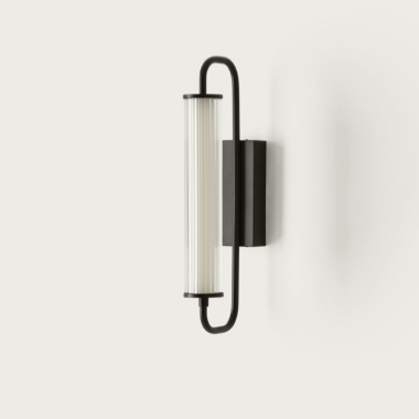 La lámpara de pared Ison, con su forma cilíndrica vertical, marco de metal negro y difusor de vidrio esmerilado acanalado, agrega un toque de elegancia industrial cuando se monta en una pared blanca.