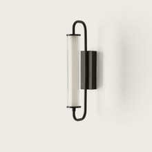  Un aplique de pared moderno que presenta una luz cilíndrica vertical de vidrio esmerilado encerrada en un marco elíptico negro, montada en una pared blanca. Esta lámpara de pared Ison combina elegancia con funcionalidad industrial.