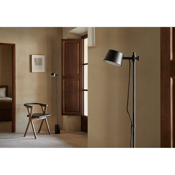 Una sala interior protagonizada por una silla de madera con un cojín negro y una elegante Lámpara de pie Nera con tecnología LED integrada, junto a una ventana con contraventanas de madera. Hay obras de arte minimalistas montadas en la pared, lo que contribuye al ambiente sereno.