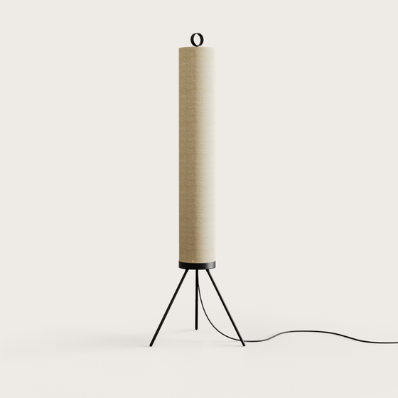 En el suelo hay una lámpara de pie cilíndrica de color beige, conocida como Lámpara de pie Nooi, con una elegante base de trípode en negro mate y un cable de alimentación.