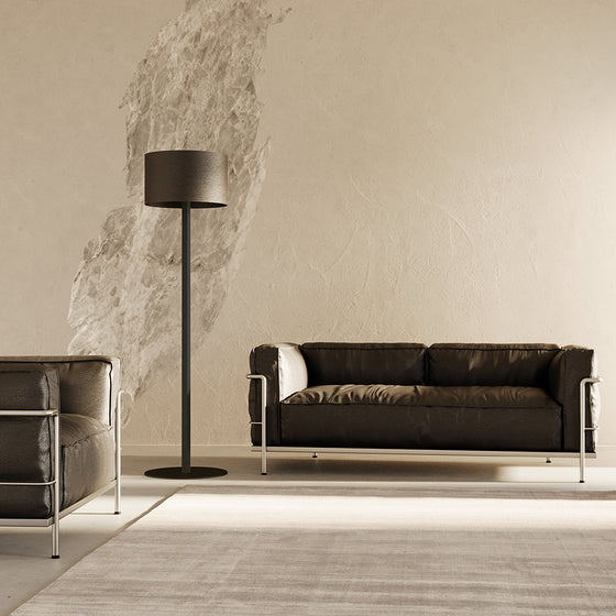 Sala de estar minimalista con un sofá de cuero marrón oscuro, un sillón a juego, una lámpara de pie Rems y arte abstracto en una pared beige texturizada.