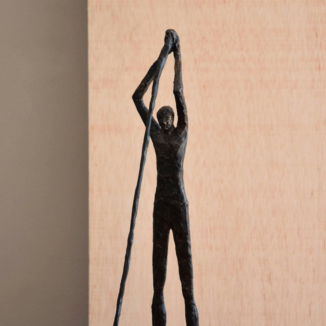La figura decorativa de metal en bronce envejecido Hombre N1 presenta una figura esbelta que se coloca con gracia y se extiende hacia arriba en un diseño minimalista sobre un fondo simple.