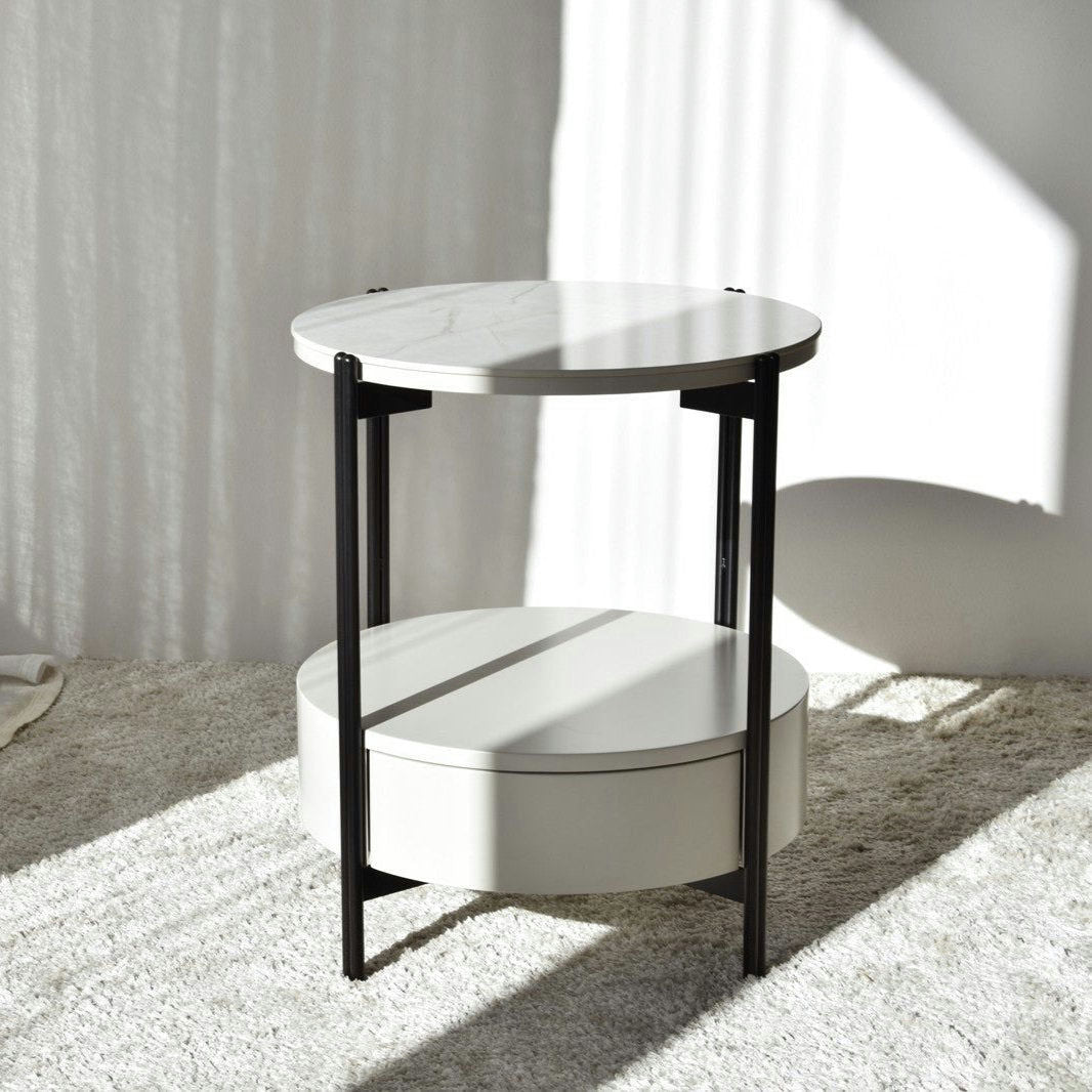 La Mesa Auxiliar N.16 redonda blanca con estante inferior y patas de metal negro, de elegante factura, adorna una alfombra clara en una habitación luminosa. Inspirada en la estética de Mármol Cerámico Blanca, realza sin esfuerzo cualquier espacio.
