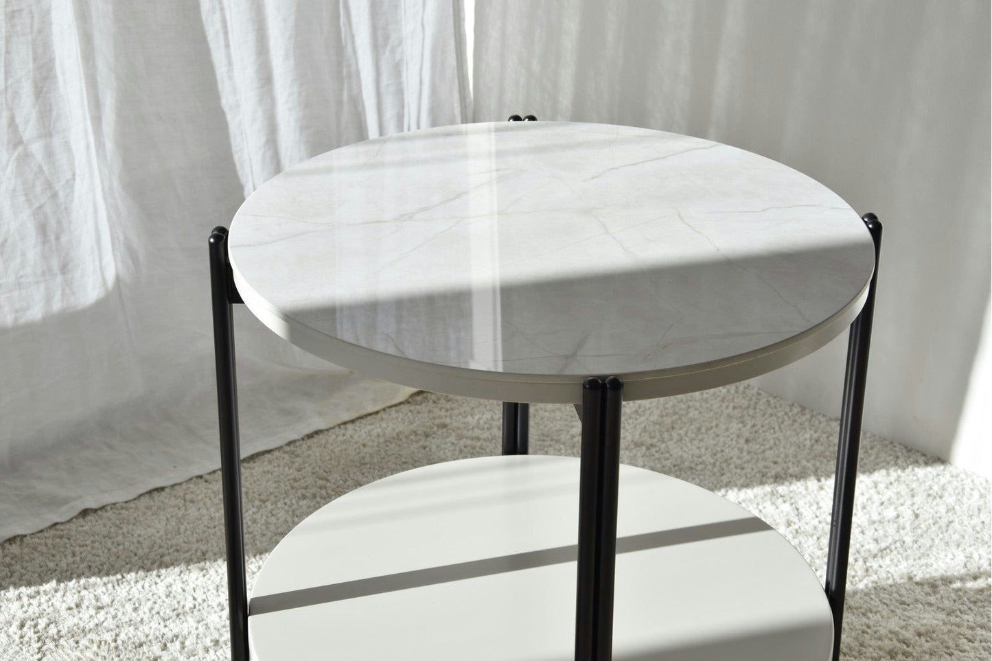 La Mesa Auxiliar N.16 con cubierta de mármol y patas de metal negro reposa sobre una alfombra clara, destilando diseño elegante mientras adorna el espacio frente a cortinas blancas.