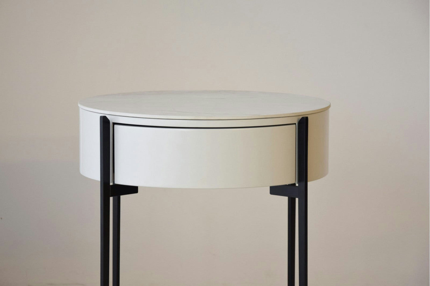 La Mesa Auxiliar N.18 es una mesa auxiliar redonda de color blanco con elegante mármol cerámico, un solo cajón y elegantes patas de metal negro sobre un fondo liso, ideal para realzar el diseño de interiores.