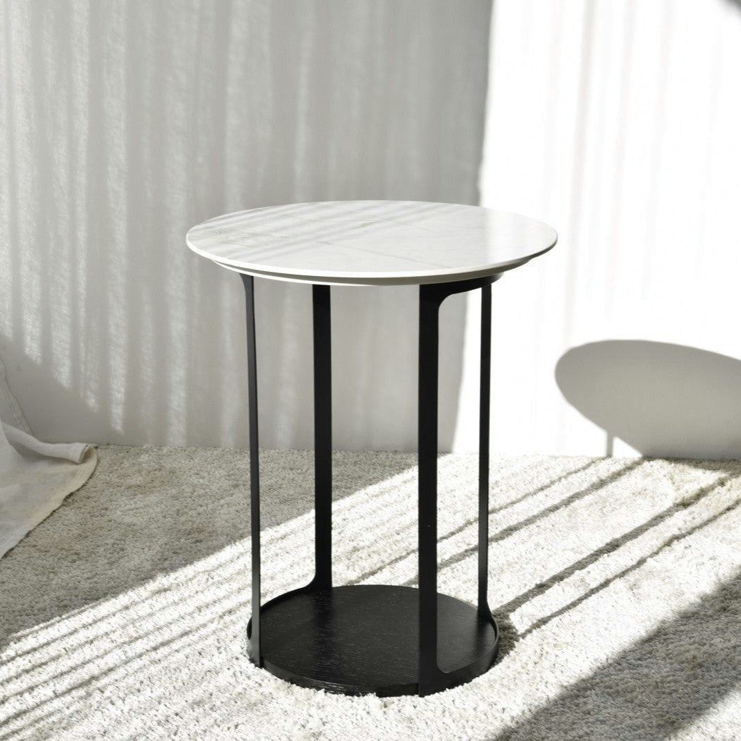 La Mesa Auxiliar N.30 Blanco y Negro de Urbicus presenta un diseño redondo moderno con una tapa blanca y patas de metal negro, que encarna el diseño contemporáneo, colocado sobre una alfombra de color claro en una habitación iluminada por el sol.