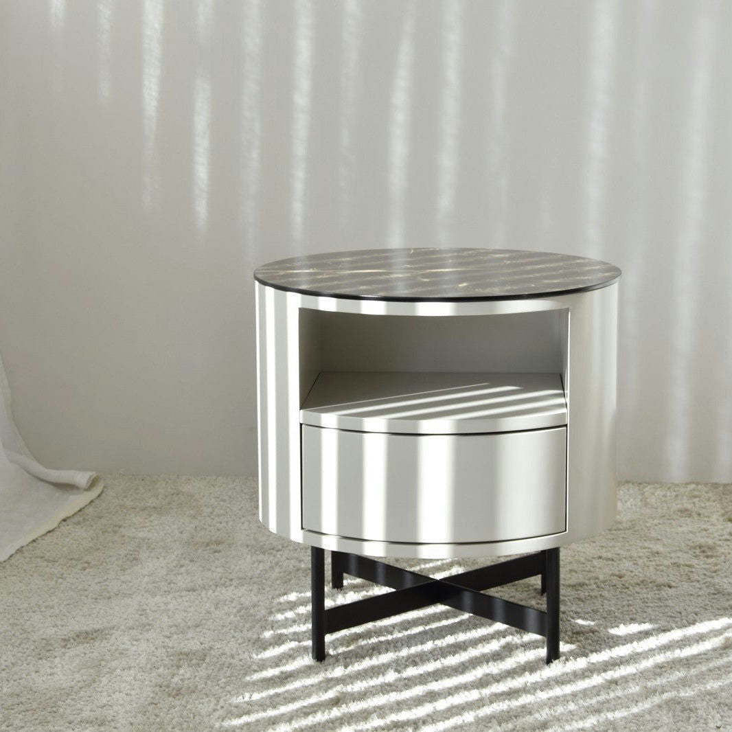 Presentamos nuestra Mesa Auxiliar N.55 - Mármol Cerámico - Gris, una elegante mesa auxiliar redonda con una superficie oscura, un estante abierto y un cajón. Ubicada contra una pared neutra, realza la decoración de interiores mientras la luz del sol proyecta sombras estampadas sobre la mesa y el piso alfombrado.