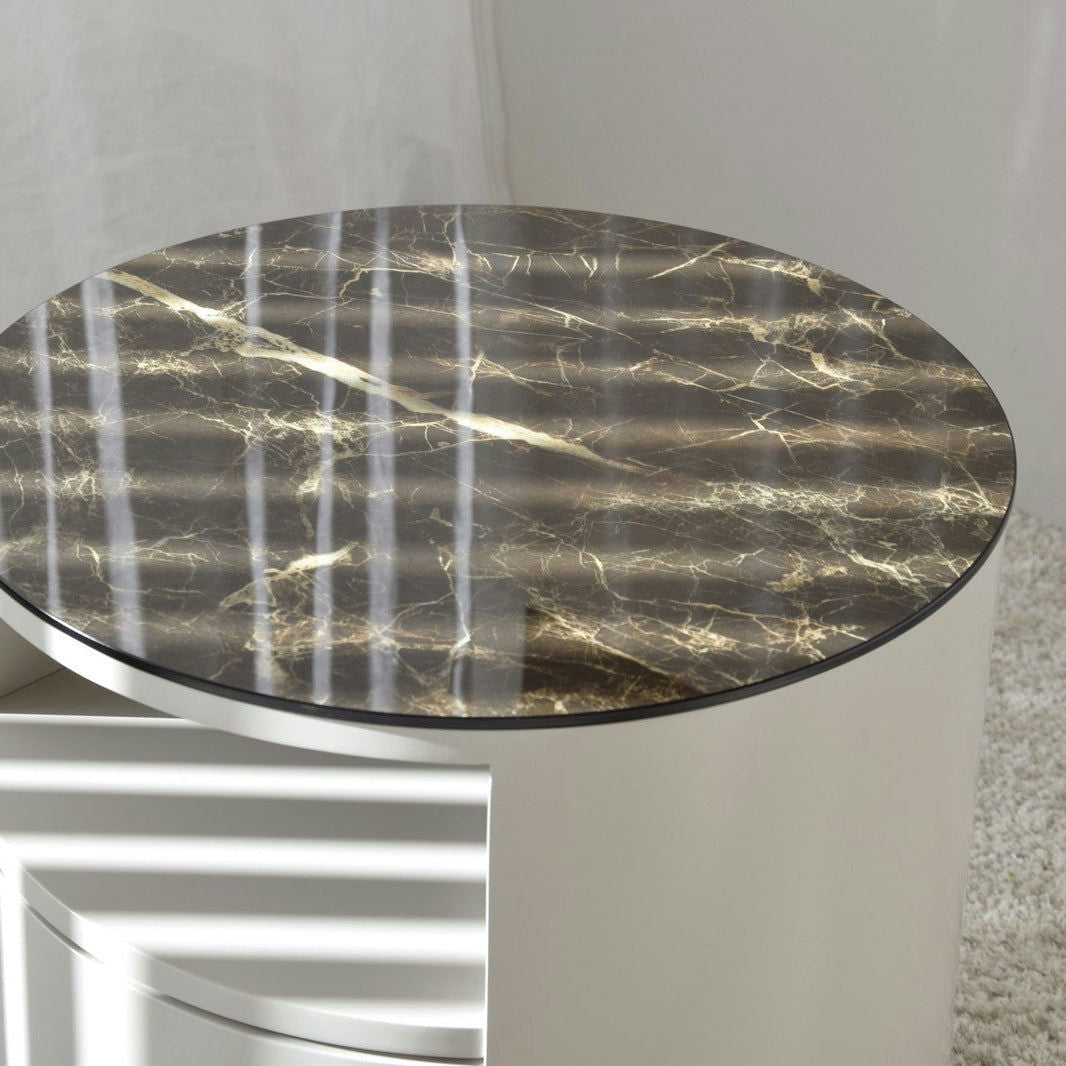 La Mesa Auxiliar N.55 es una mesa llamativa con una superficie de cerámica de mármol brillante con patrones veteados en marrón oscuro y una base blanca curva, que representa un diseño moderno. La luz del sol proyecta sombras rayadas sobre esta elegante pieza de mármol cerámico.