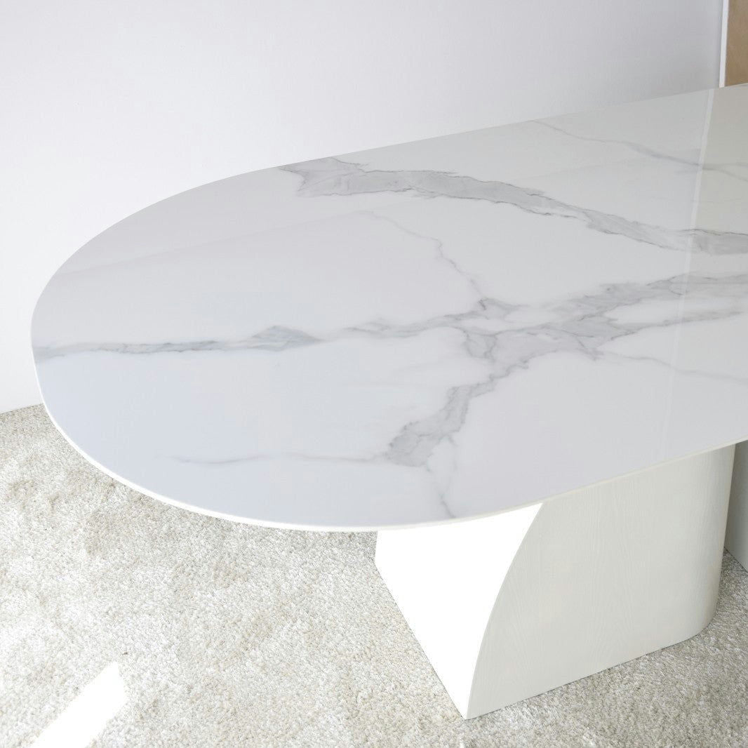 La Mesa de Comedor Blanco-Gris, de forma ovalada. Norte muestra elegantes vetas grises, que recuerdan al mármol blanco veteado, colocadas sobre una alfombra clara contra una pared blanca inmaculada.