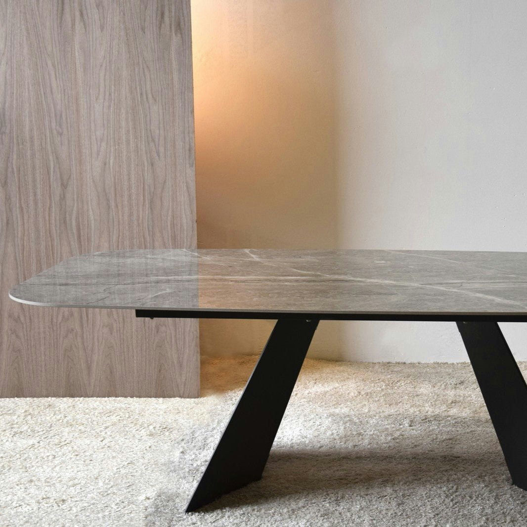 La mesa de comedor Shulján presenta una moderna tapa rectangular de color gris cerámico y una base angular de metal negro, colocadas sobre un piso alfombrado claro con un fondo de paneles de madera.