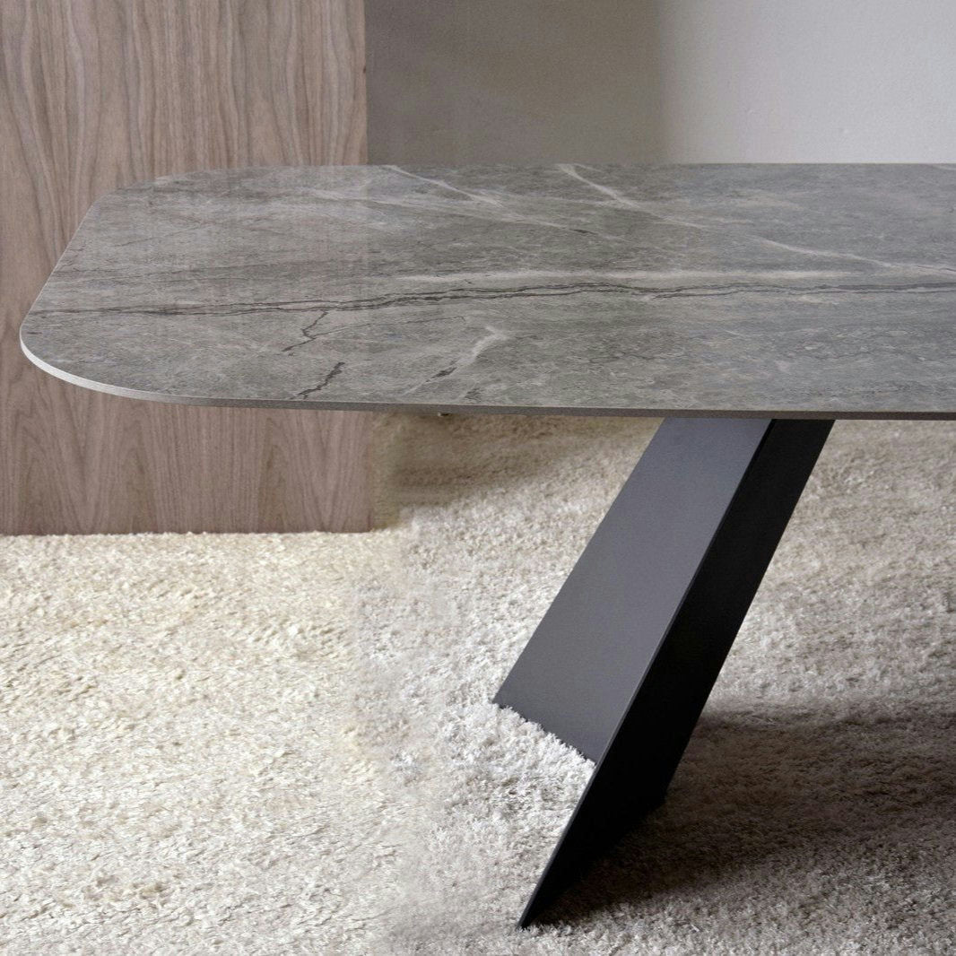 La Mesa de Comedor Cerámico Gris, Base en Metal - Shulján cuenta con un elegante diseño contemporáneo con una superficie rectangular de mármol gris sostenida por una única pata diagonal de metal negro, elegantemente presentada sobre una alfombra texturizada de color claro.