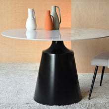  La Mesa de Comedor Cerámico Redonda - Eleganza, con su tapa blanca y base negra, muestra tres jarrones en blanco, naranja y gris. Una silla con patas negras es parcialmente visible a la derecha sobre una alfombra texturizada.