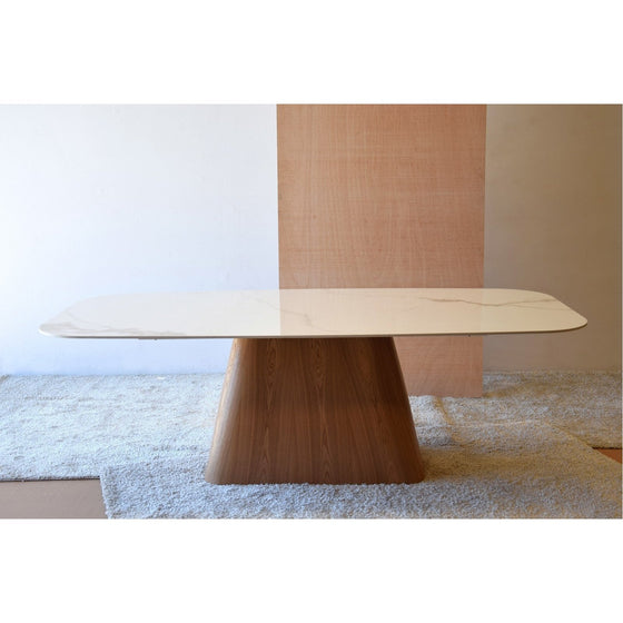 Una moderna Mesa de Comedor Cerámico con Base Fresno Mate - Marmorea rectangular se alza sobre una alfombra gris claro sobre un fondo de paneles de madera.