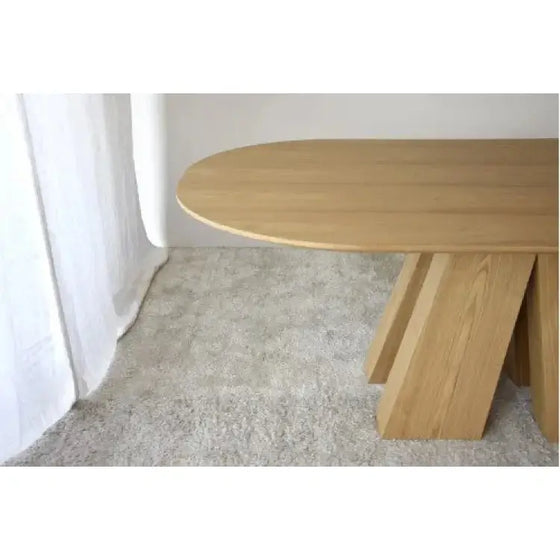 Una Mesa de Comedor Fresno Natural Elba elegante con una forma ovalada frente a una cortina blanca.