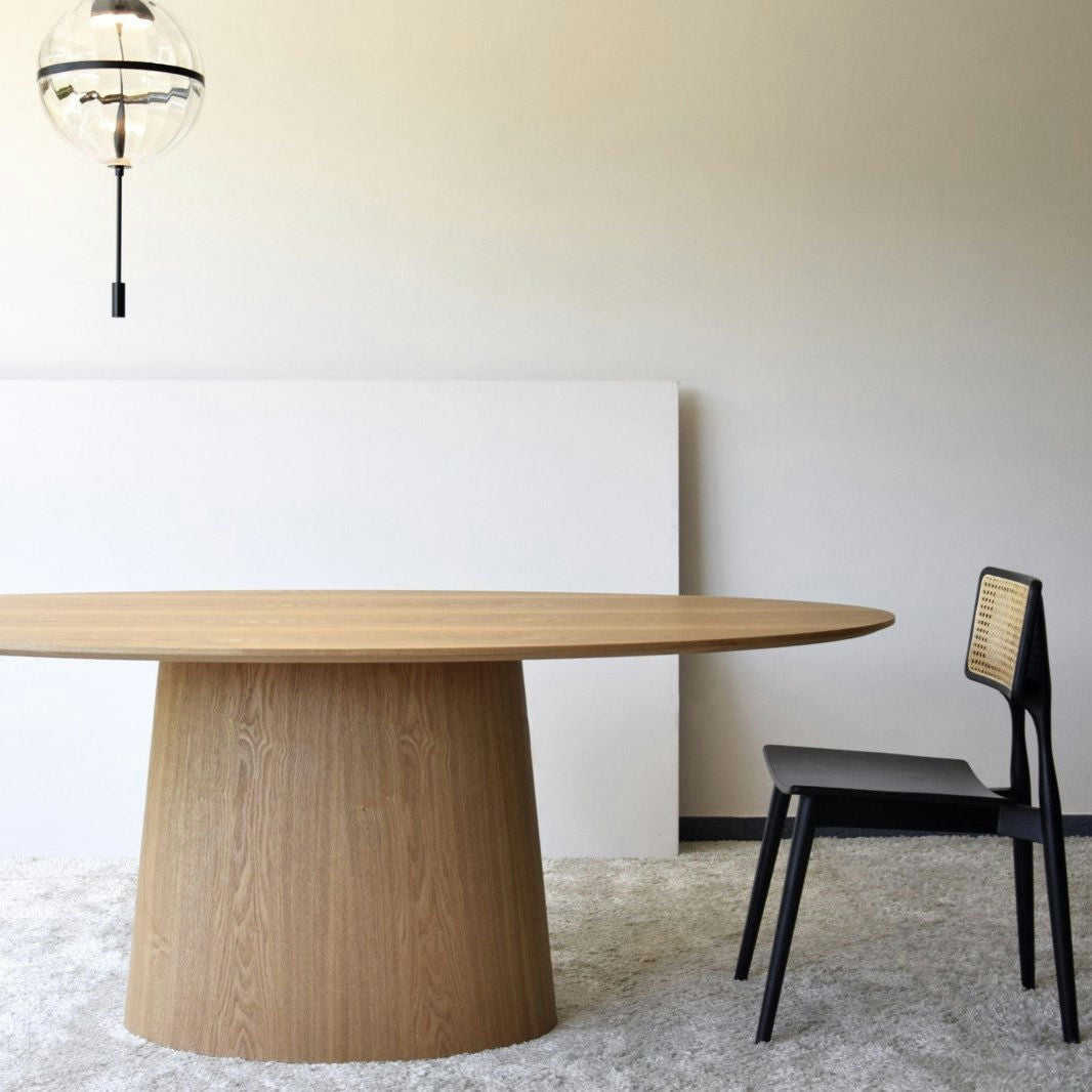 Una habitación minimalista muestra la Mesa de Comedor Oval Fresno Natural - Abies combinada con una silla negra con respaldo de caña, complementada por una luz colgante esférica del techo, que incorpora un elegante diseño de interiores.