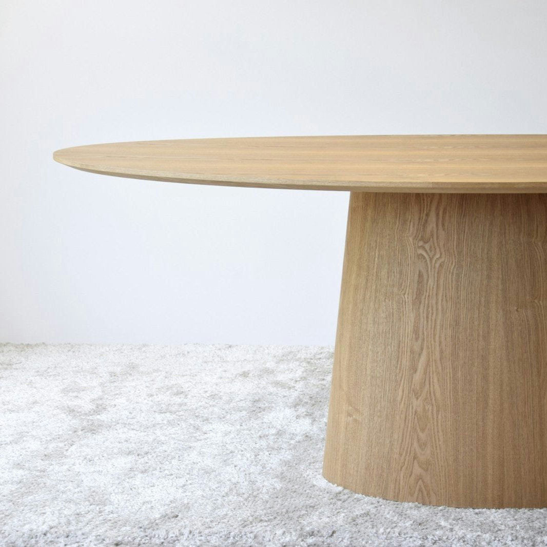 Una Mesa de Comedor Ovalada Fresno Natural - Abies con una base cilíndrica gruesa se encuentra sobre una lujosa alfombra de color claro en una habitación minimalista, lo que refleja un exquisito diseño de interiores.