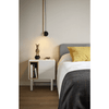 Un dormitorio minimalista con una Mesita Corvo 2C blanca con libros y una pequeña estatua, al lado de una cama con cabecero gris y almohada amarilla.