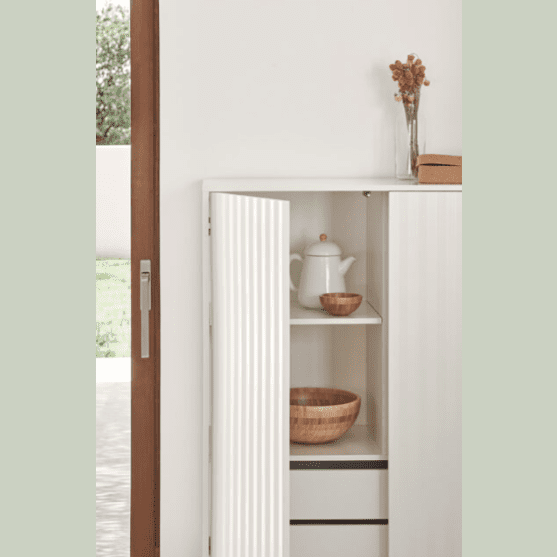 Un rincón de cocina minimalista con un Mueble Auxiliar Sierra 2P4C blanco que muestra una tetera y un cuenco de madera sobre estantes, visible a través de una puerta parcialmente abierta.