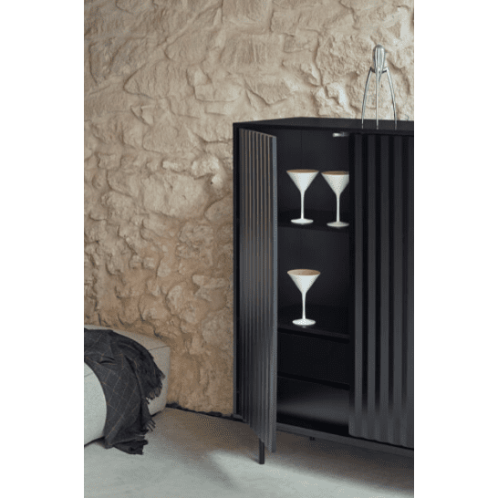 Un moderno Mueble Auxiliar Sierra 2P4C negro que muestra tres elegantes copas de martini, colocadas contra una pared de piedra texturizada en una habitación con poca luz y diseño minimalista.