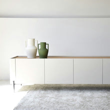  El Mueble TV Arena Mate y Roble Natural - Armonízate presenta un diseño minimalista con una cubierta de roble natural y un acabado lacado blanco mate, mostrando elegantemente dos grandes jarrones en blanco y verde sobre una alfombra gris claro, agregando una sutil sofisticación a su espacio vital.