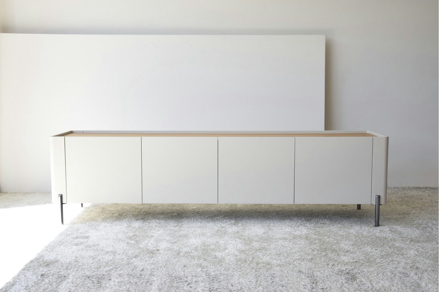 El Mueble TV Arena Mate y Roble Natural - Armonízate, con un diseño blanco minimalista y elegantes patas de metal, se encuentra sobre una alfombra gris texturizada en una habitación sencilla.