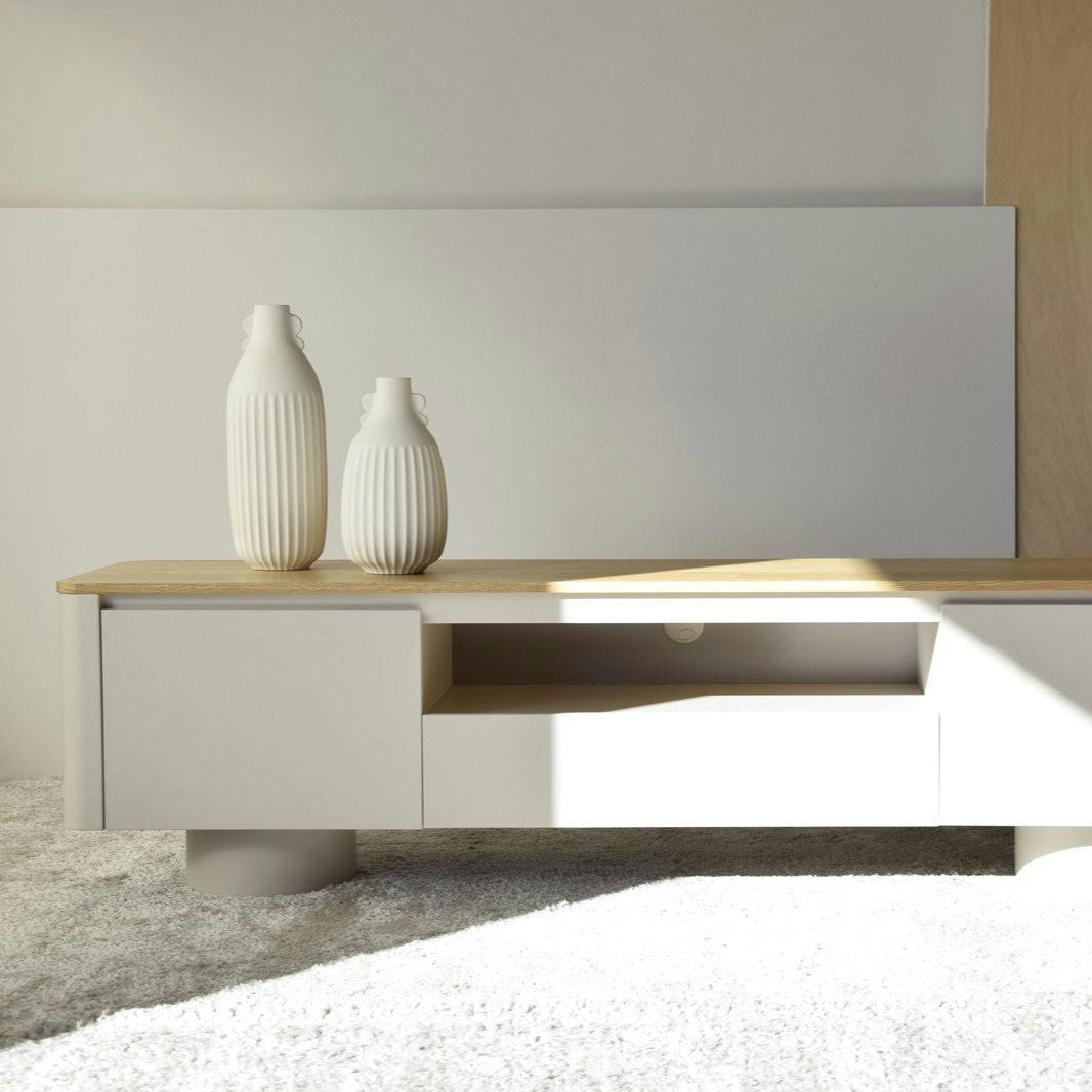Una habitación minimalista cuenta con un banco bajo de madera blanca con dos jarrones blancos grandes, patrones que reflejan la luz del sol en el banco y piso alfombrado. La atmósfera serena se ve realzada por la sutil elegancia de un Mueble TV Gris Arena y Roble Cilindro que se integra a la perfección con el diseño.