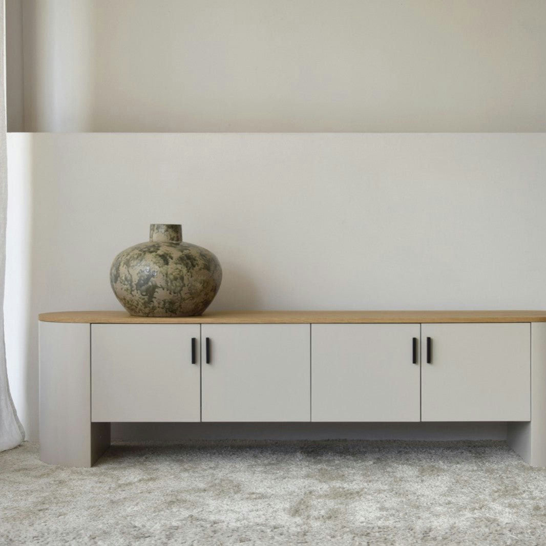 Una habitación minimalista exhibe un gran jarrón de cerámica en el Mueble TV Gris Arena y Roble - Tea, con un acabado gris claro con una tapa de roble natural y manijas negras, sobre una pared gris arena con piso alfombrado.