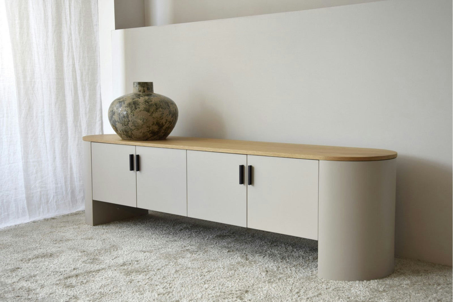 El Mueble TV Gris Arena y Roble - Tea tiene bordes redondeados, una tapa de roble natural y manijas negras. Sobre él reposa un gran jarrón decorativo. Se complementa con una cortina blanca y una alfombra de color claro en el fondo sereno, que realza su elegante acabado gris arena.