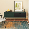 Un Mueble TV Punto 2P2C de color verde oscuro con almacenamiento oculto, un póster enmarcado encima y elementos decorativos encima, colocado contra una pared beige sobre una alfombra multicolor.